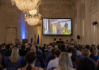 In einem Saal mit prachtvollen Kronleuchtern und kunstvollen Stuckwänden sitzen festlich gekleidete Menschen und schauen auf eine Videoleinwand.