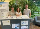 Drei Menschen stehen lächelnd an einem Informationsstand mit Broschüren zum Studium am Fachbereich Rechtspflege.